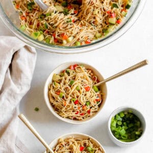 Cold sesame noodles served in a white bowl with a gold fork on a white background. Small bowl of green onions off to the side.