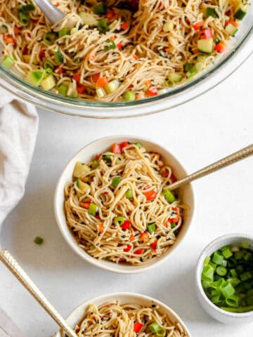 Cold sesame noodles served in a white bowl with a gold fork on a white background. Small bowl of green onions off to the side.
