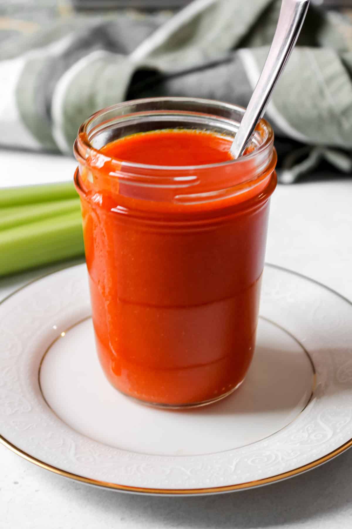 Buffalo sauce in a glass jar with a spoon in the jar. Jar is on a white plate.