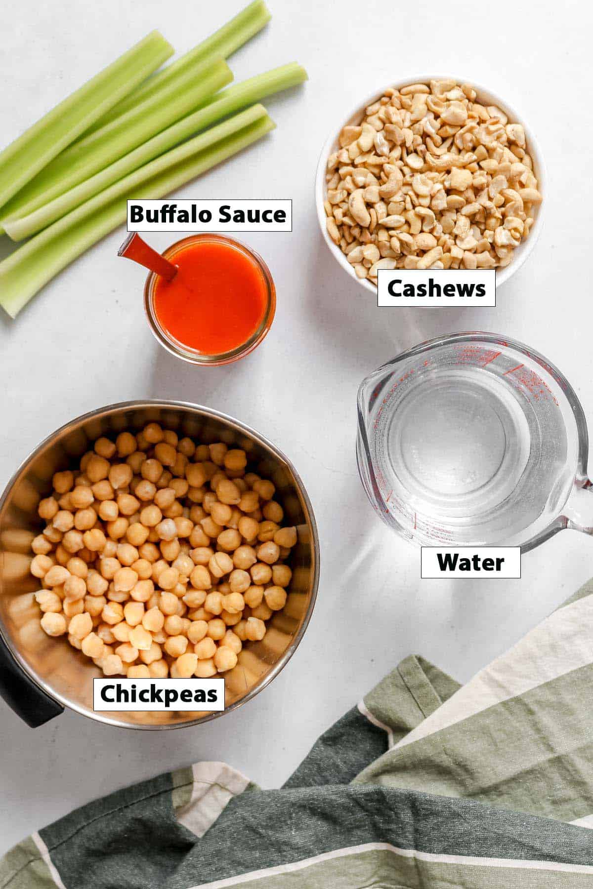 Individual ingredients to make buffalo dip in bowls and labeled: Chickpeas, buffalo sauce, water, and cashews