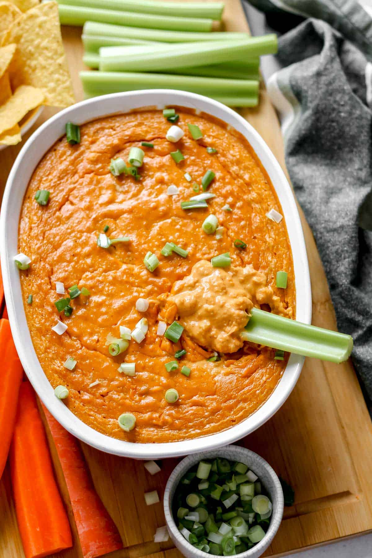 Plant-based buffalo chicken dip topped with green onions in a white baking dish, surrounded by celery, carrots, and tortilla chips, with celery dipped into the dip.
