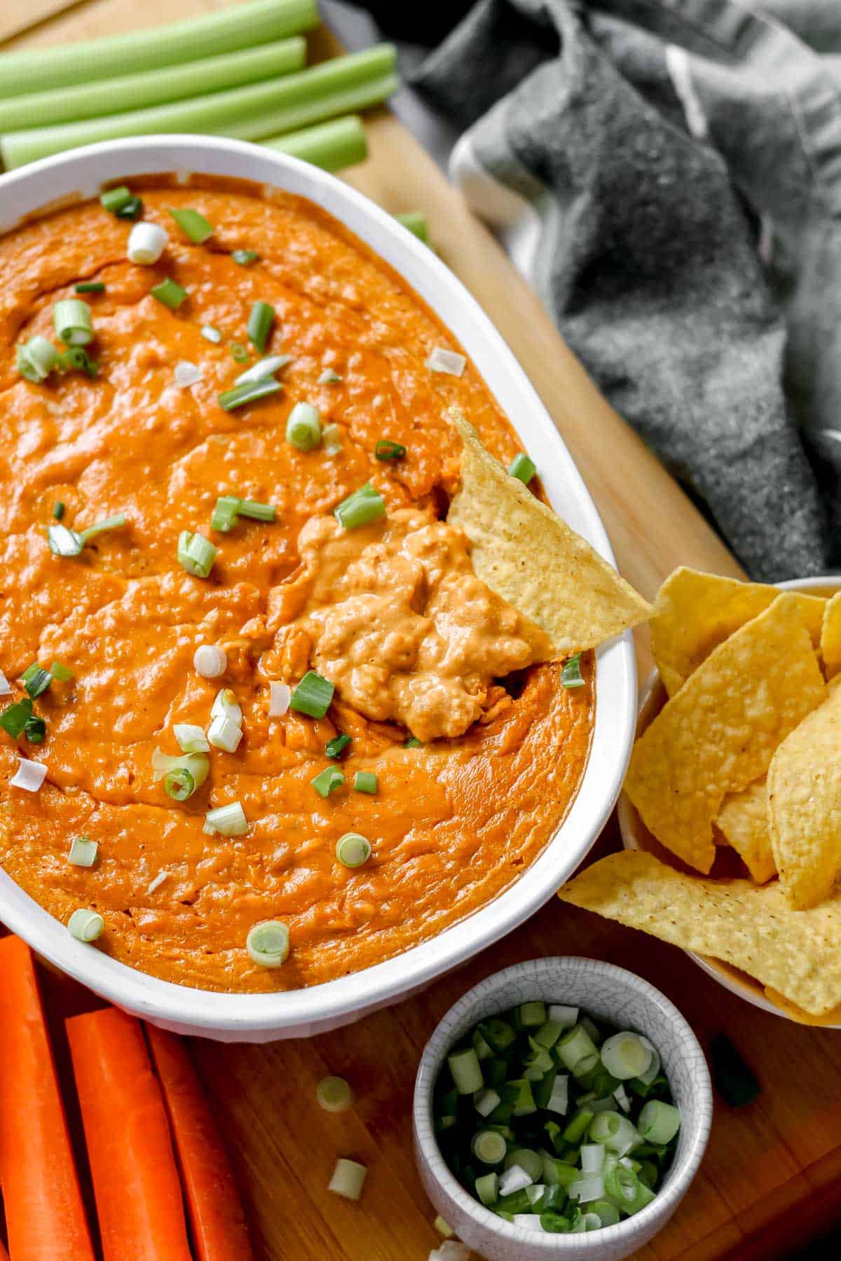 Vegan buffalo chicken dip topped with green onions in a white baking dish, surrounded by celery, carrots, and tortilla chips, with a chip dipped into the dip.