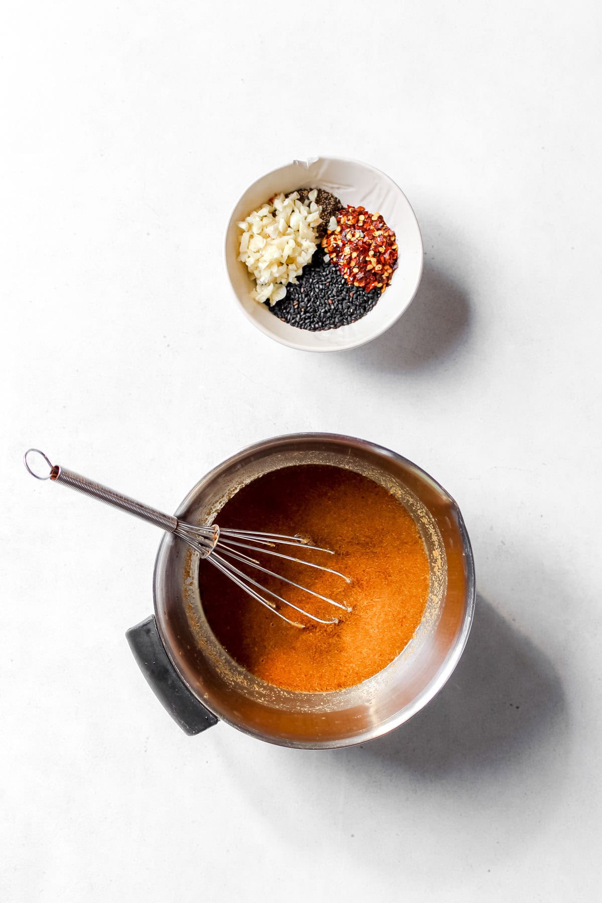 Small white bowl filled with sesame seeds, chopped garlic, red pepper flakes, and black pepper and a stainless steel bowl filled with the sauce and a whisk.