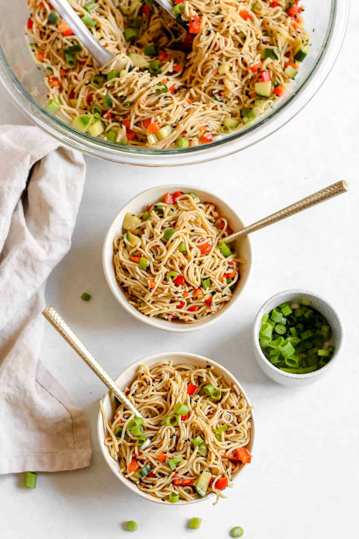 Cold sesame noodles served in two white bowls with gold forks on a white background. Small bowl of green onions off to the side.
