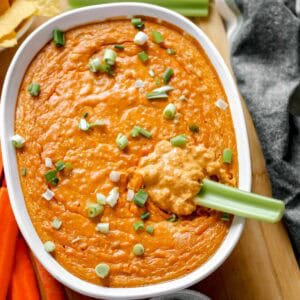 Vegan Buffalo Chicken Dip topped with green onions in white baking dish on cutting board surrounded by celery, carrots, and chips.