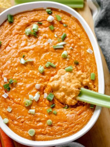 Vegan Buffalo Chicken Dip topped with green onions in white baking dish on cutting board surrounded by celery, carrots, and chips.