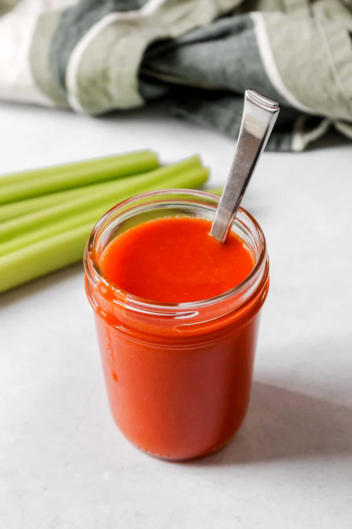 Vegan buffalo sauce in glass container along with a spoon. Jar is next to celery sticks and there is a green towel in the background.