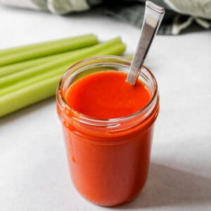 Buffalo sauce in a glass jar with a spoon in the jar. Celery is next to the jar.