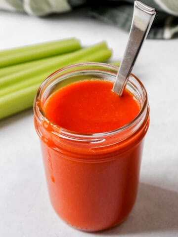 Buffalo sauce in a glass jar with a spoon in the jar. Celery is next to the jar.