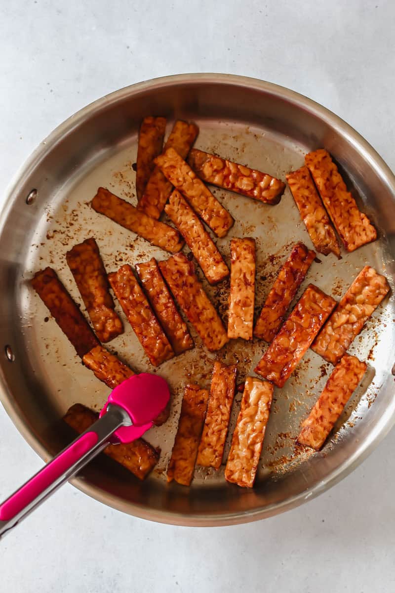 Cooking tempeh bacon in stainless steel skillet with pink tongs