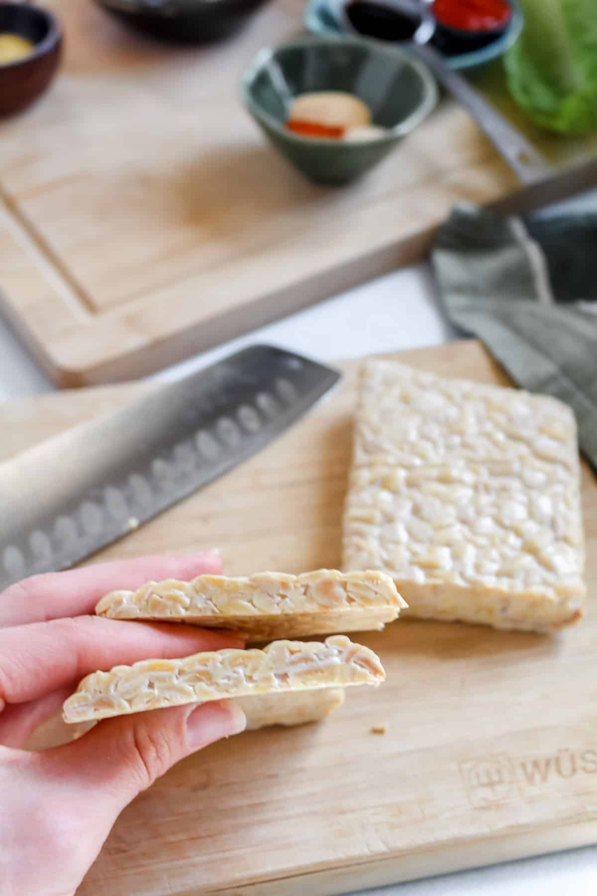Tempeh sliced lengthwise into a thin slice being held by a hand over a wooden cutting board