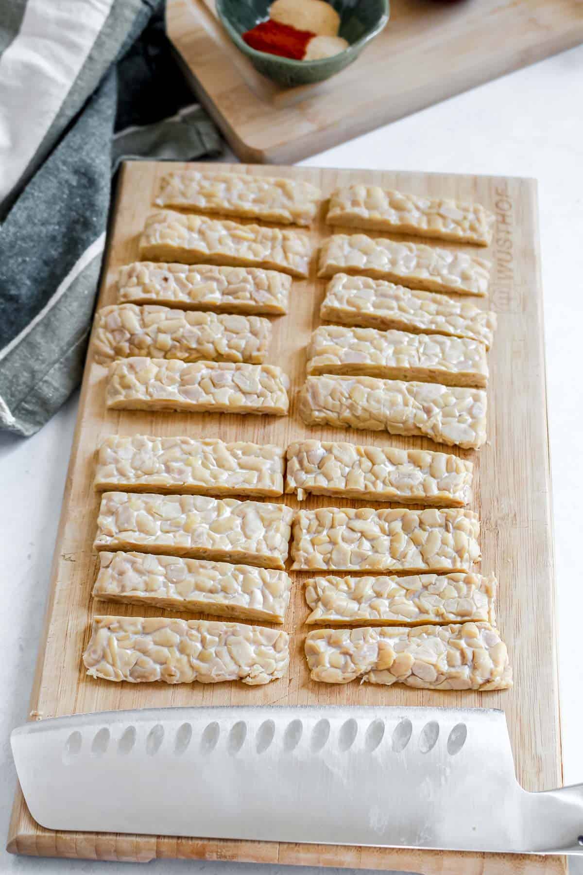 Tempeh sliced into thin strips on a wooden cutting board with a knife
