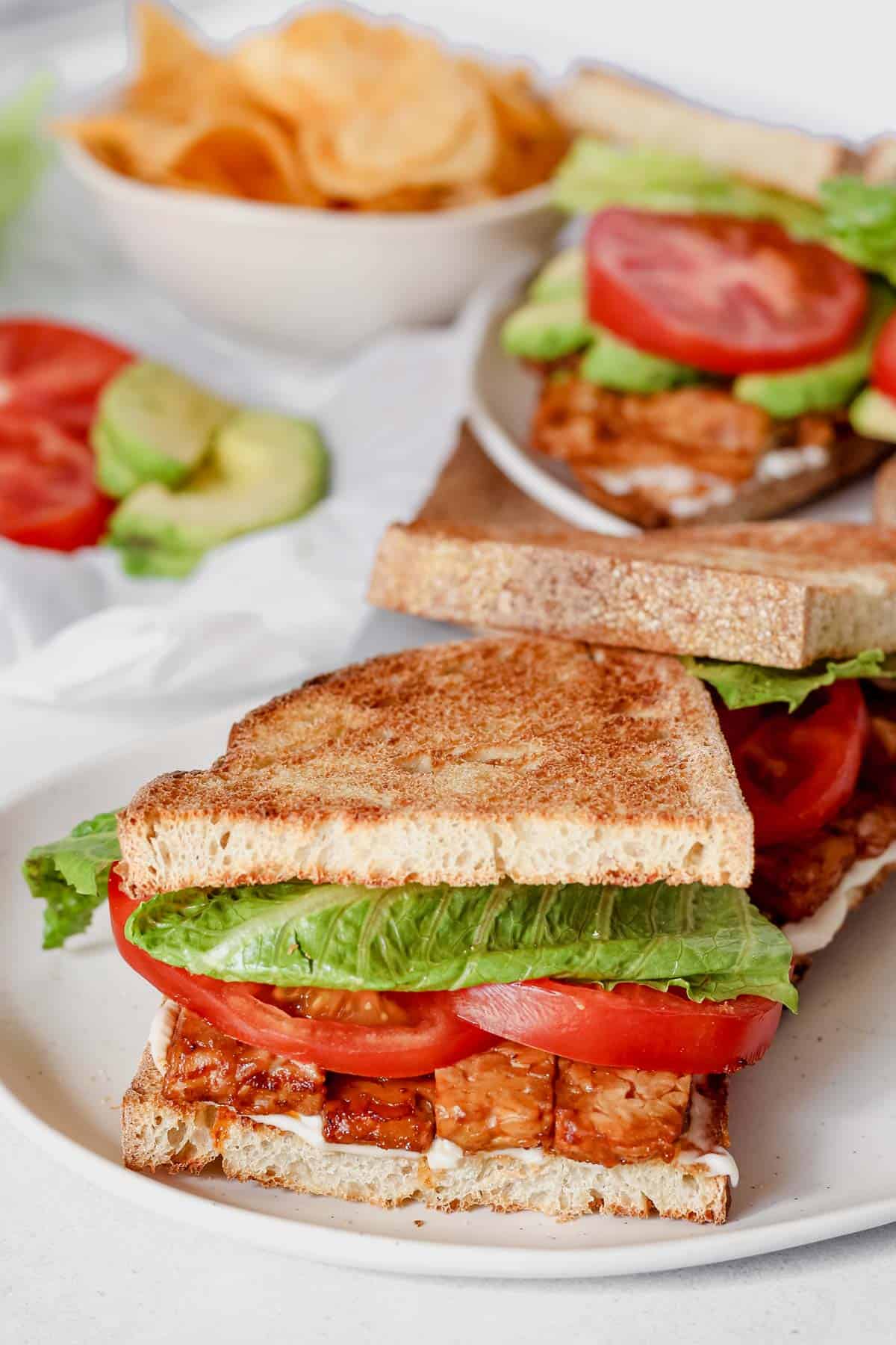 Close up of sandwich made with toasted bread, tempeh bacon, lettuce, tomato and vegan mayonnaise on a white plate