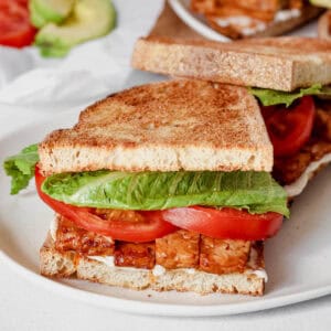 Sandwich containing tempeh bacon, lettuce, tomato and vegan mayonnaise on a white plate